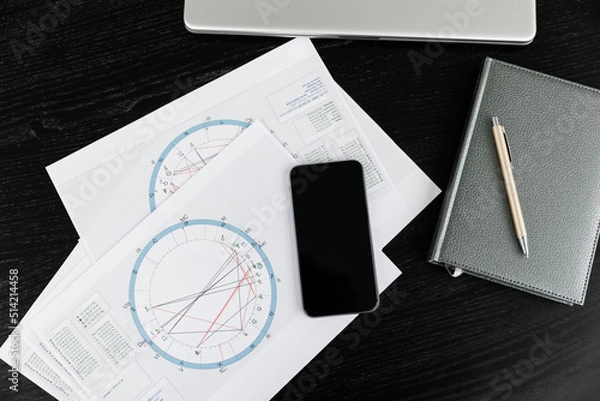 Fototapeta The notepad for writing down ideas and the astrological natal birth chart lie on a black background. A computer keyboard and mouse next to it and a phone. Astrological consultation idea.