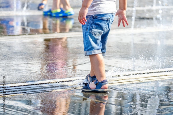 Fototapeta The feet of a child in sandals play with the jets of the fountain in the square. Selective focus. The city is very hot.