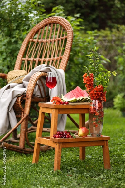 Fototapeta Summer garden. Rocking chair, straw hat, green trees. Bouquet with red flowers, fresh watermelon, pear and cherry on the table, stool. A glass of red wine. Summer atmosphere. Background image