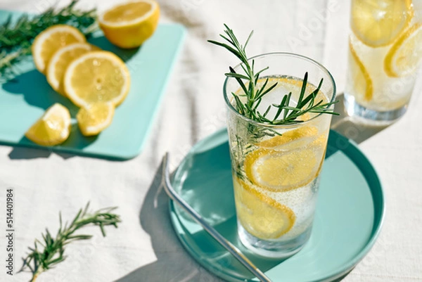 Fototapeta Letnie orzeźwiający napój lemoniadowy lub koktajl alkoholowy z lodem, rozmarynem i plasterkami cytrynowymi na stole w ogrodzie. Świeży, zdrowy zimny napój cytrynowy. Woda z cytryną.