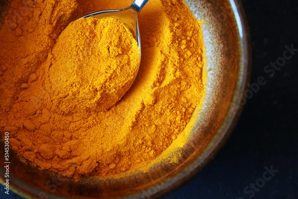 Fototapeta close-up of yellow turmeric spice powder with a spoon in a  brown ceramic bowl on dark background, top view of turmeric powder as cooking spice