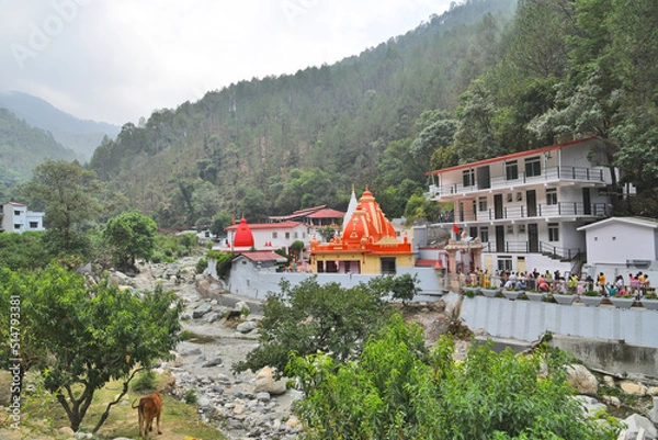 Obraz Kainchi Dham Temple, Uttarakhand, where a river flows next to a sacred temple that is surrounded by hills and trees.