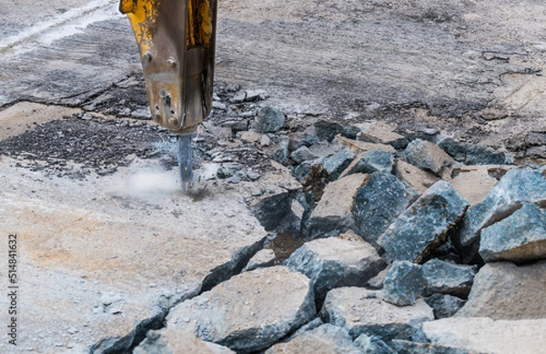 Fototapeta Close-up of demolition hammer with dust and sparks and broken concrete pieces. Detail of hydraulic jackhammer mounted on excavator arm working at old road reconstruction. Repairs in civil engineering.