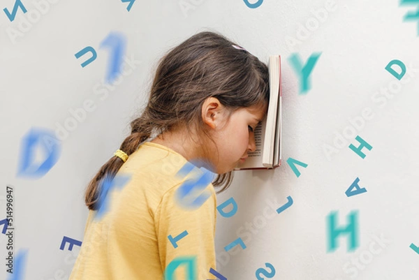 Fototapeta Sad and tired caucasian girl with dyslexia holds a book on her forehead. Flying tangled letters in the air. The child learns to speak and read correctly
