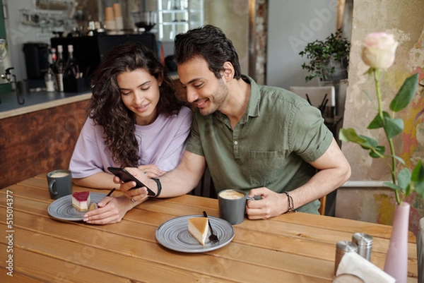 Fototapeta Young couple communicating in video chat in smartphone while sitting by table in cafe and having cappuccino or latte with tasty cheesecakes
