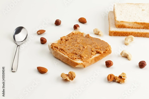 Obraz Square bread for toast with peanut butter. Nuts, spoon, bread slices and a peanut butter sandwich on a white table.