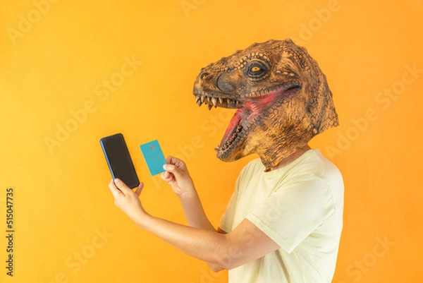 Fototapeta Happy unrecognizable person wearing dinosaur animal head mask , holding smartphone and credit card on orange background