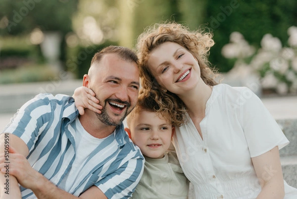 Obraz Father, mother, and son are sitting on the steps in the garden of an old European town. A close photo of a laughing family in the park in summer at sunset.
