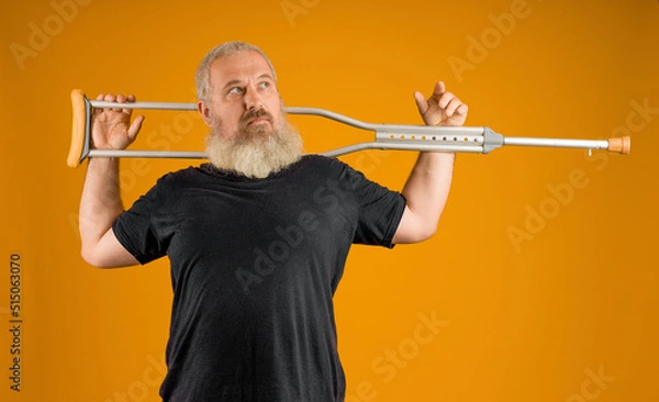 Fototapeta disabled man with a gray beard put a crutch on his shoulders on a yellow background