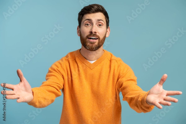 Fototapeta Come to me! Portrait of friendly kind man holding arms wide open for free hugs and smiling happily, welcoming, going to embrace. Indoor studio shot isolated on blue background
