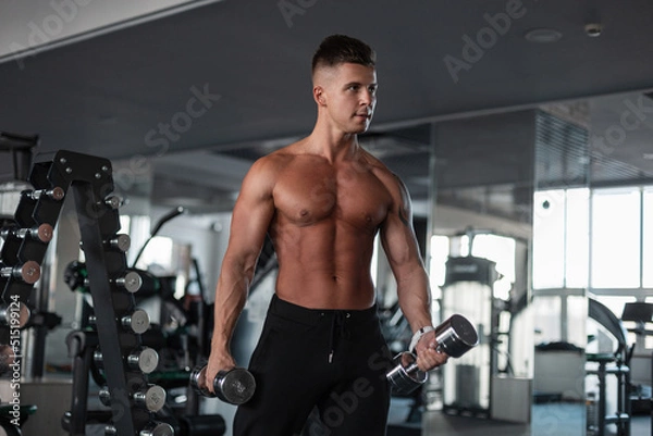 Fototapeta Cool handsome young male bodybuilder athlete with a sporty muscular naked torso working out in the gym with dumbbells