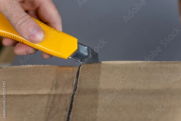 Fototapeta A man's hand opens a cardboard box with a yellow cutter. The flaps of the package are sealed with adhesive tape. Close-up. Selective focus.