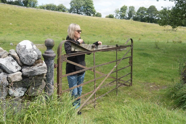 Fototapeta An attractive blonde lady leans and rests on an old iron farm gate in rural setting with traditional dry stone wall.She wears sunglasses and is smiling.Yorkshire