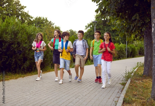 Obraz Back to school. Cheerful group of junior high school students with backpacks on their shoulders walk on sidewalk in park. Joyful children in summer casual clothes go and talk to each other on warm day