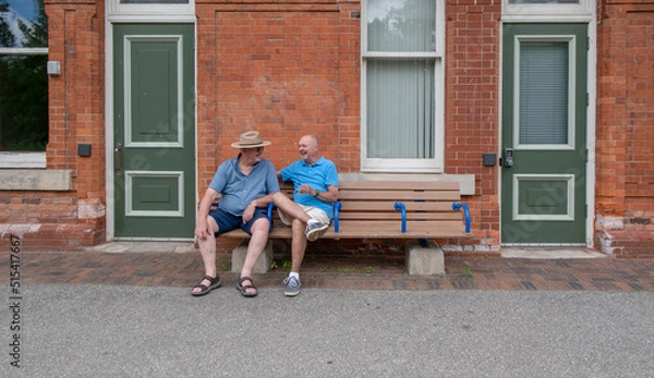 Fototapeta An older gay married couple in light conversation, sit next to each other on a an outdoor train station bench on a sunny summer's day.