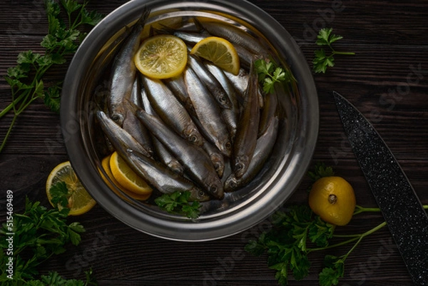 Fototapeta In a metal plate, which stands on a dark wooden table, there is a lot of small fish. Greens and lemons lie next to the plate.