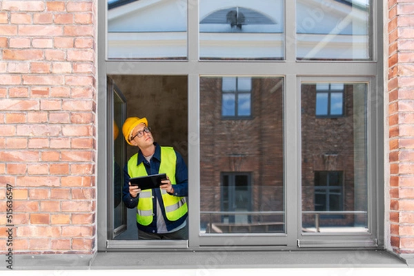 Fototapeta architecture, construction business and building concept - male architect in helmet and safety west with tablet pc computer in window