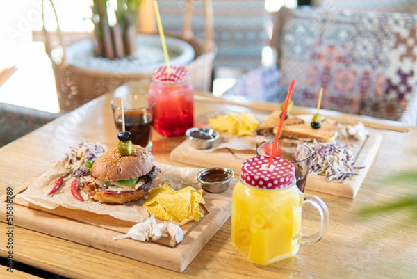 Obraz Beef patty burger, bacon, cheese, tomato, greens, pickle, goose pate sandwich, red cabbage salad, hot pepper, fries, corn chips, ketchup, orange and cherry juice