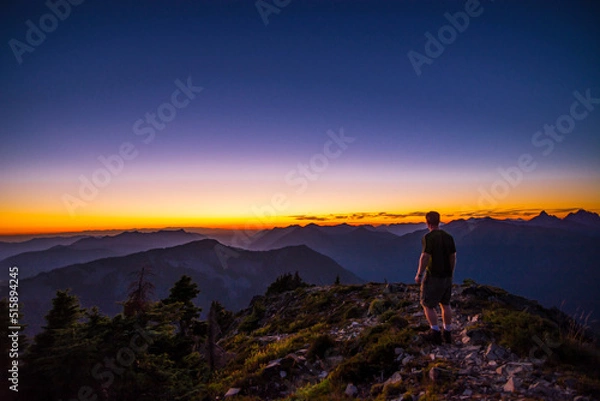 Fototapeta An athletic adventurous male hiker standing on top of a mountain looking out at a beautiful sunset.