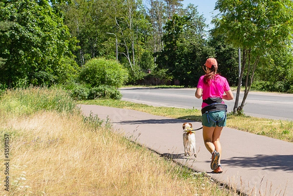 Fototapeta A female athlete in a pink T-shirt, shorts and a red baseball cap runs with a dog along the track. Canicross. Sports running with a dog. Lifestyle.