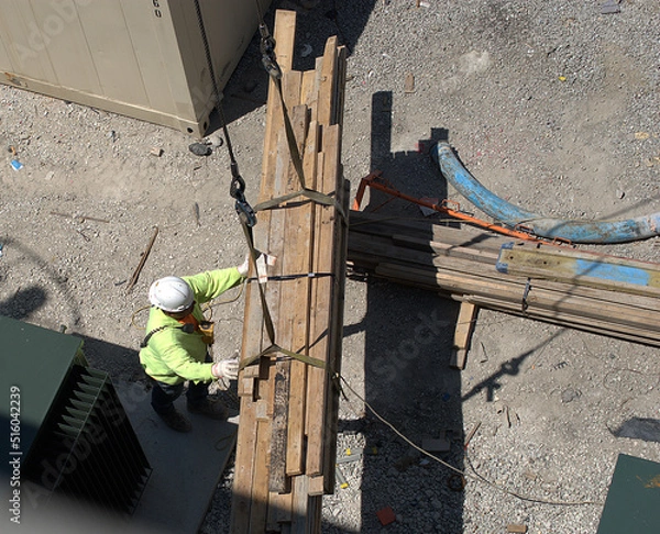 Obraz Chicago, Illinois, USA. June 28, 2022. Looking down on a construction worker who is operating a remote controled crane to move a bundle of lumber.