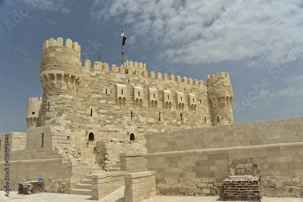 Fototapeta Citadel of Qaitbay, Alexandria, Egypt with the Egyptian flag on top and a background of blue cloudy sky. The Fort of Qaitbay is a 15th-century defensive fortress located on the Mediterranean sea coast