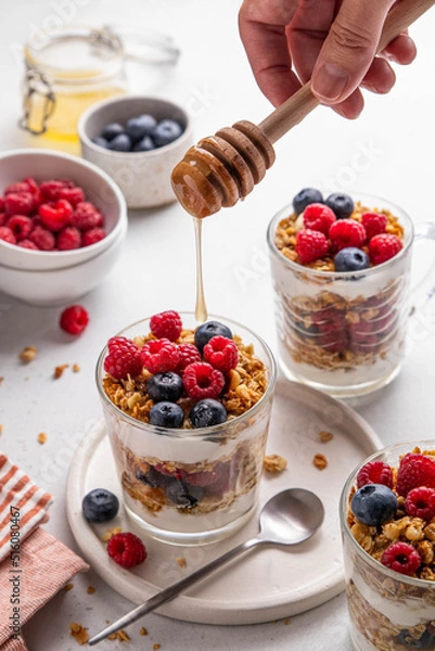 Fototapeta Granola with yogurt in glass served with fresh raspberry, blueberry and honey. Homemade oatmeal muesli. Process of pouring honey on granola. Honey drips from wooden spoon in hand. Selective focus.