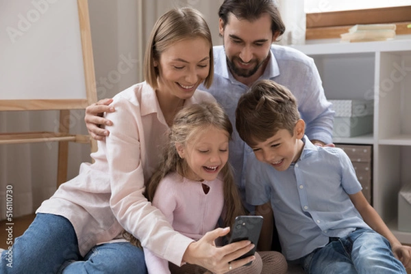 Fototapeta Happy joyful couple of parents and two little kids making video call on mobile phone, chatting together, taking family selfie, resting on warm floor at home, looking at screen, webcam, laughing