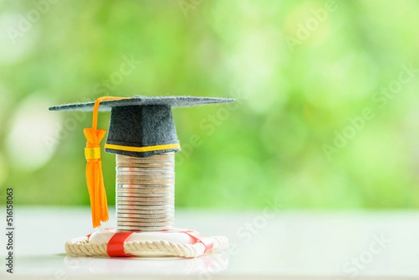 Fototapeta Tuition protection service and tuition refund insurance, financial concept : Black graduation cap or a mortarboard placed higher on top of a coin stack with a red lifebuoy on a table, green background