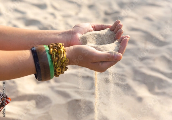 Fototapeta Female hands with many boho bracelets, and sand in the beach.