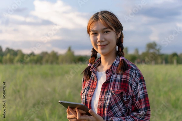 Obraz Young agriculture student working with tablet in field at sunset. Young attractive Asian girl in a field using tablet. Portrait of female farm worker with digital tablet