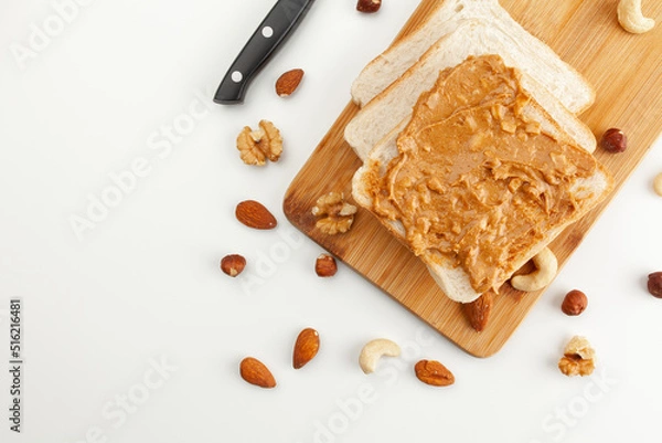 Obraz Square bread for toast with peanut butter on a wooden board. Nuts, a knife and a wooden cutting board with a sandwich and slices of bread on a white table.