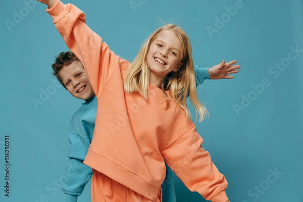 Fototapeta two cute children of school age in multi-colored sweaters stand on a blue background, a boy and a girl stand with their hands apart, depicting airplanes. The theme of friendship between children