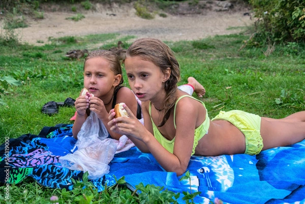 Fototapeta Two little girls on a green lawn eat a bun