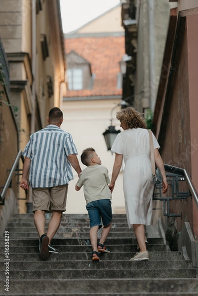 Obraz A full-length photo from the back of a family which is climbing stairs in an old European town. A happy father, mother, and son are holding hands and having fun in the evening.