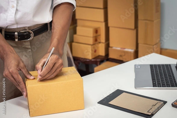 Fototapeta Small business owner packing in the cardbox at workplace. Cropped shot of man preparing a parcel for delivery at online selling business office. Ecommerce drop shipping shipment service concept.