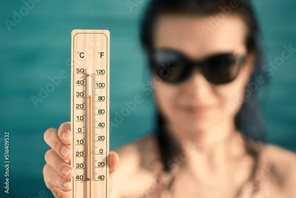 Fototapeta Beautiful young woman in sunglasses with a thermometer in her hand on the background of the sea. Summer vacation and heat concept, selective focus
