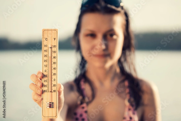 Fototapeta Beautiful young woman with a thermometer in her hand on the background of the lake. Summer vacation and heat concept, selective focus