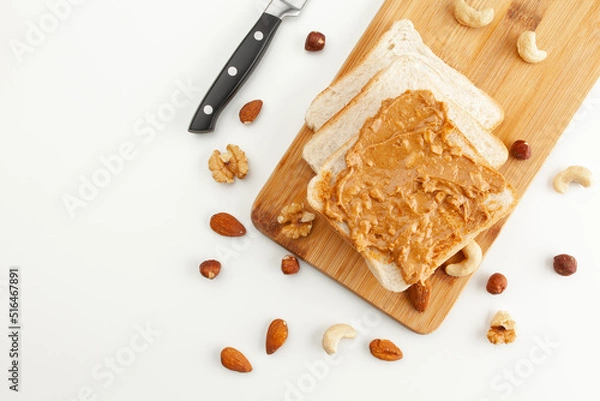 Obraz Square bread for toast with peanut butter on a wooden board. Nuts, a knife and a wooden cutting board with a sandwich and slices of bread on a white table.
