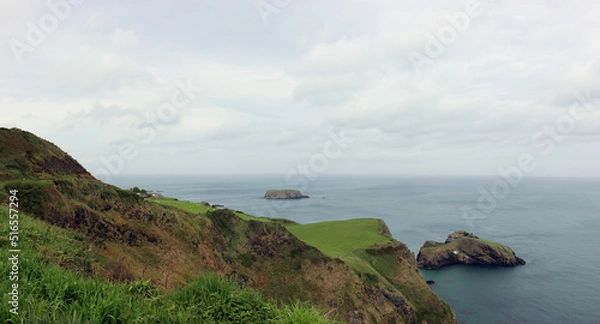Fototapeta Cloudy weather on the green slopes of the coast of Northern Ireland over the idyllic island of Carrick-a-Rede with its narrow suspension bridge