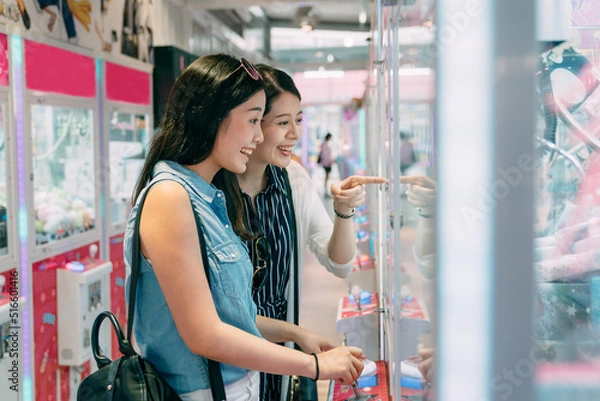 Fototapeta side portrait two happy female friends are looking with excitement and pointing the toy in the showcase while having fun playing claw machine in a small store.