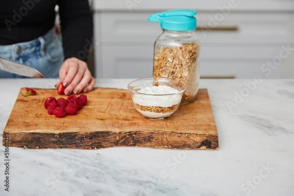 Obraz Yogurt with granola, and raspberries and strawberries next to jar with granola in a bright modern kitchen and a woman cutting strawberries