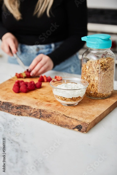 Obraz Yogurt with granola, and raspberries and strawberries next to jar with granola in a bright modern kitchen and a woman cutting strawberries