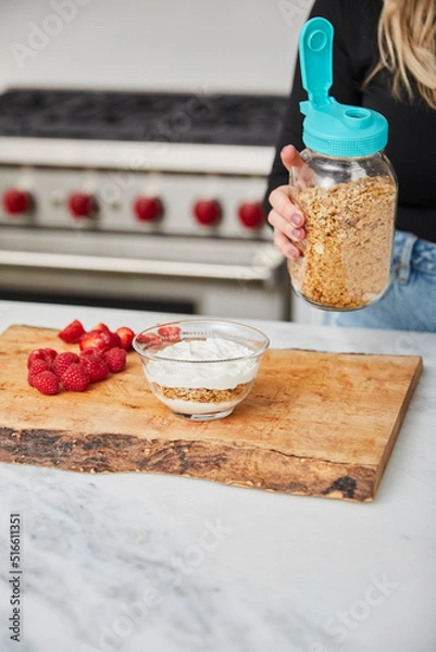 Obraz Yogurt with granola, and raspberries and strawberries and a female holding jar with granola in a bright modern kitchen