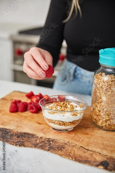 Obraz A woman topping yogurt and granola with fruit in a bright modern kitchen
