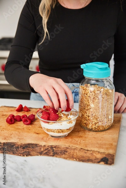 Obraz A woman topping yogurt and granola with fruit in a bright modern kitchen