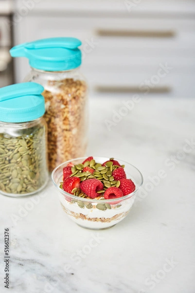 Obraz Yogurt with granola topped with raspberries and strawberries next to jars with granola and nuts in a bright modern kitchen