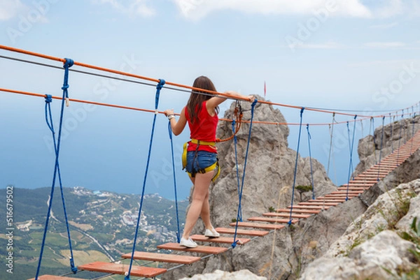 Fototapeta A young girl walks across a suspension bridge on an abyss in the mountains against the backdrop of the sea on a sunny summer day.Safe adventures while traveling, healthy image zhini concept