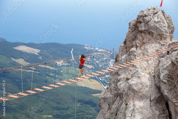 Fototapeta A young girl walks across a suspension bridge on an abyss in the mountains against the backdrop of the sea on a sunny summer day.Safe adventures while traveling, healthy image zhini concept