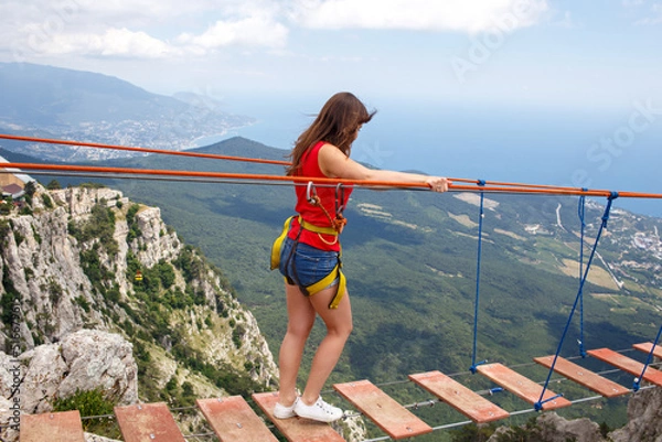 Fototapeta A young girl walks across a suspension bridge on an abyss in the mountains against the backdrop of the sea on a sunny summer day.Safe adventures while traveling, healthy image zhini concept